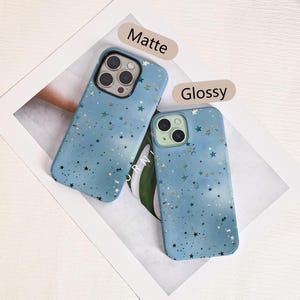 May include: Two blue phone cases with a star pattern. One case has a matte finish, and the other has a glossy finish. The cases are lying on a white surface with a green leaf visible.