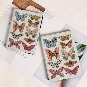 Puede incluir: Dos fundas rectangulares para lectores electrónicos con diseño de mariposas. Las fundas son de color blanquecino con borde negro y presentan varias ilustraciones de mariposas coloridas. Una funda tiene el nombre "Gemma" impreso.