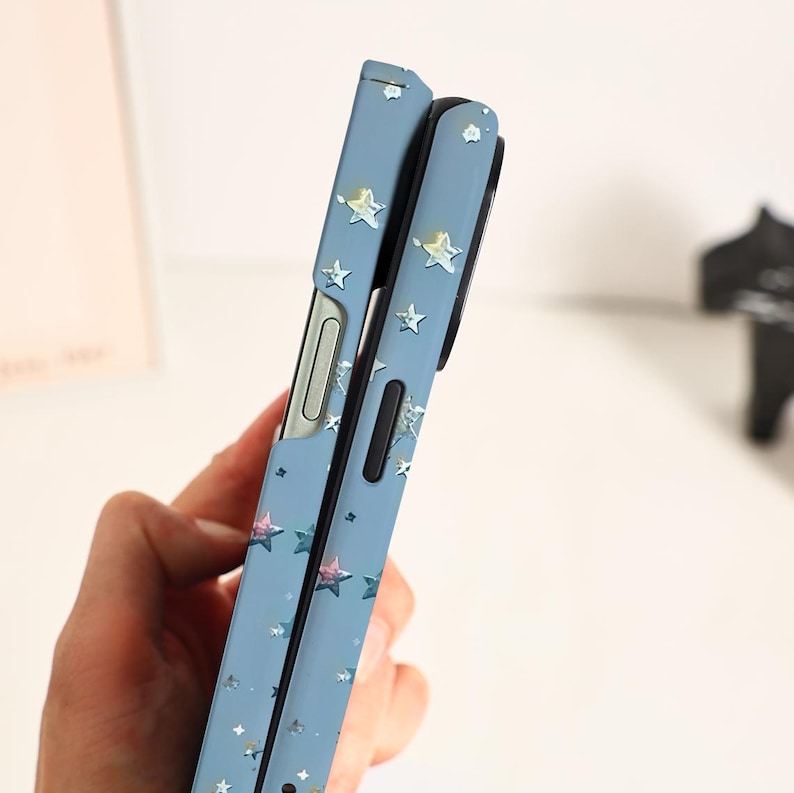 May include: A light blue phone case with a star pattern. The case has a folding design and features silver and pink star accents. The phone case is designed to protect the phone from scratches and damage.