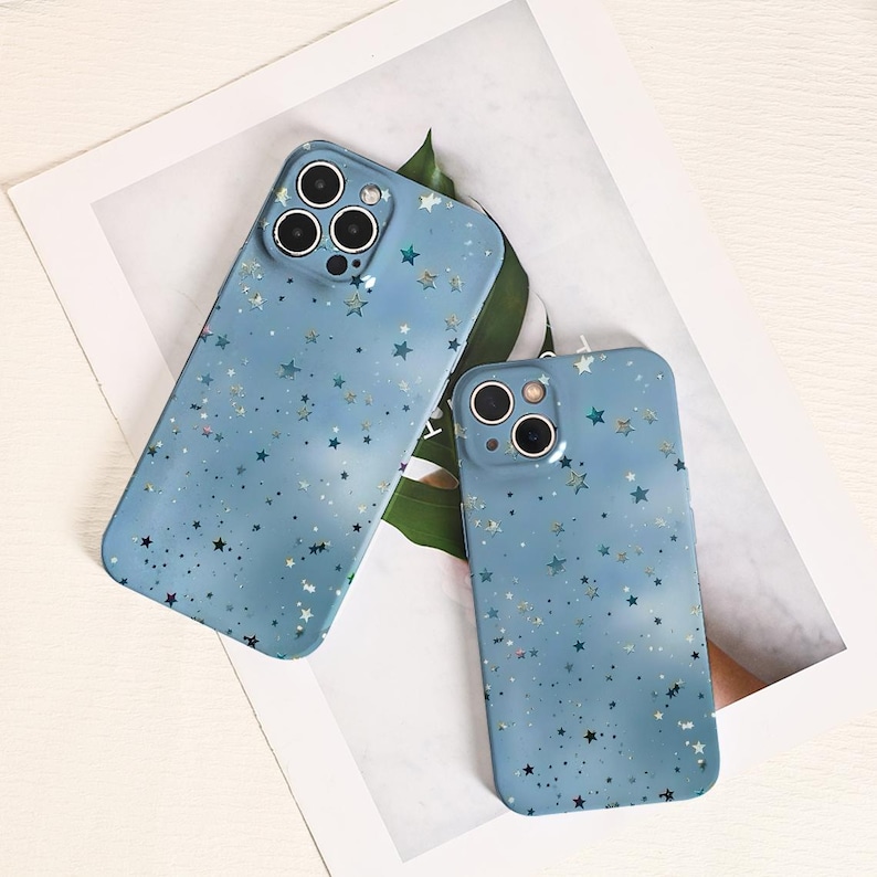 May include: Two light blue phone cases with a star pattern. The cases have a matte finish and feature a scattering of small, silver and dark blue stars. The cases are designed to fit a smartphone and have cutouts for the camera.
