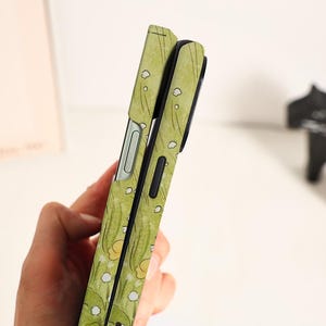 May include: A green phone case with a floral pattern featuring yellow and white flowers and green leaves. The case has a sleek design with a black border and a silver-colored button. The case is designed to protect the phone.