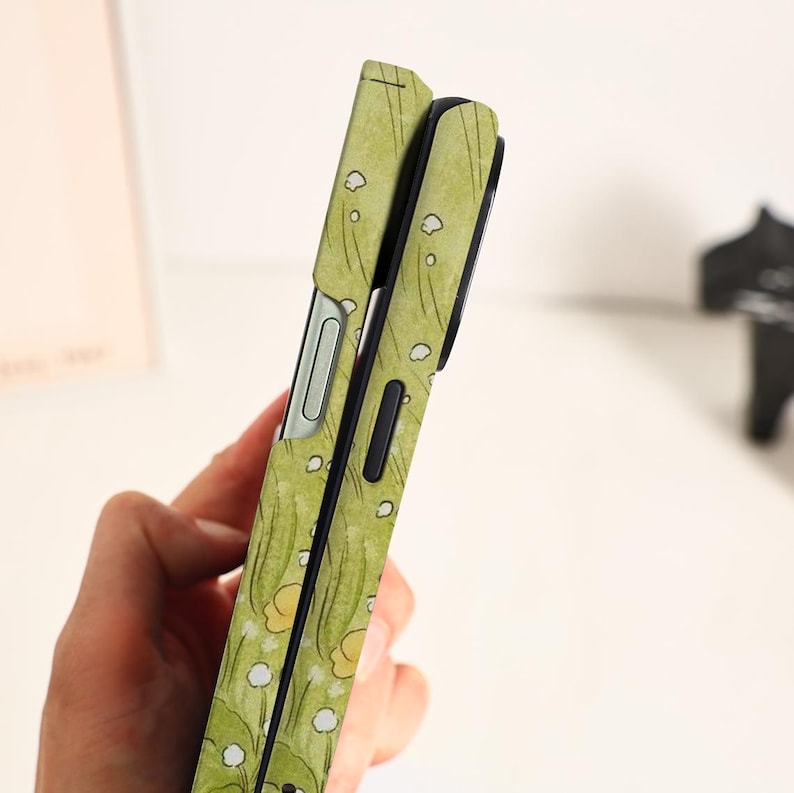 May include: A green phone case with a floral pattern featuring yellow and white flowers and green leaves. The case has a sleek design with a black border and a silver-colored button. The case is designed to protect the phone.