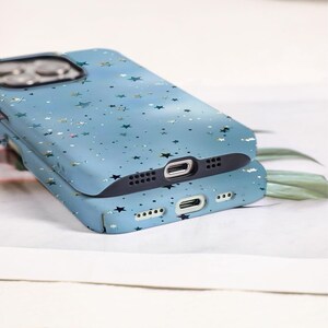 May include: Two light blue phone cases stacked on top of each other. The cases are decorated with a pattern of various sized stars and specks. The cases have black edges and openings for the camera, charging port, and speakers.