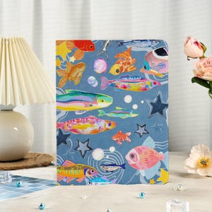 Puede incluir: Un cuaderno con una cubierta azul que presenta una colorida ilustración de varios peces, estrellas y burbujas. Los peces son en tonos naranja, rosa, amarillo y azul. El cuaderno tiene una espina redondeada.
