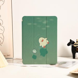 Puede incluir: Una funda para tablet verde con una ilustración de dibujos animados de una rana y un pájaro sobre un fondo verde.