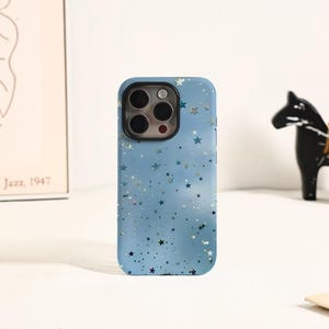 May include: A light blue phone case with a star pattern. The case has a glossy finish and features a cluster of stars in various sizes and colors, including silver and gold. The phone case has a camera cutout with multiple lenses.