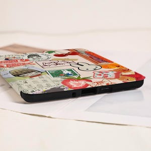 May include: A rectangular electronic device case featuring a collage of colorful illustrations including a white rabbit, red cherries, a sliced strawberry, a postage stamp, and a cartoon character with brown hair, all set against a cream background.