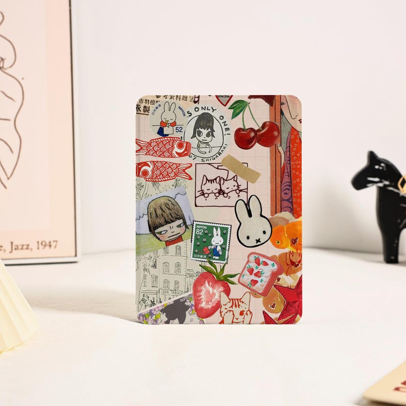 May include: A rectangular tablet case featuring a collage of various illustrations including a white rabbit, red cherries, a strawberry, a cat, and Japanese text. The design incorporates multiple stamp-like graphics and a beige adhesive tape element.