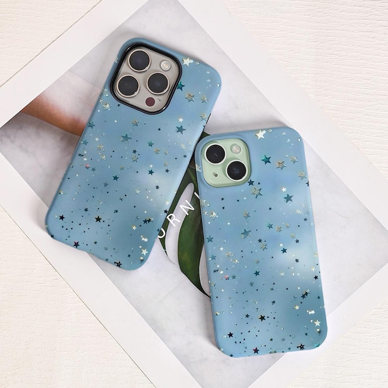 May include: Two light blue phone cases with a starry design. The cases have a matte finish and feature a pattern of various sized stars in silver and darker blue. The cases are lying on a white surface.