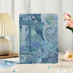 May include: A blue and white journal featuring an ocean-themed illustration. The design includes a lighthouse, whales, an octopus, starfish, and other marine life. The cover has a textured appearance and a watercolor style.