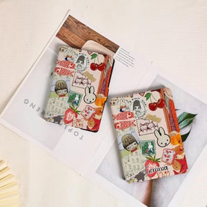 May include: Two rectangular e-reader cases featuring a collage of illustrations including a white rabbit, cherries, red fish, a person with short hair, and a green postage stamp. One case displays the name Gemma in cursive black script.