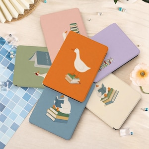 May include: A collection of six rectangular e-reader cases in various colours including orange, light blue, sage green, cream, lavender, and pink. Each case features a graphic illustration of a white goose interacting with stacks of books.