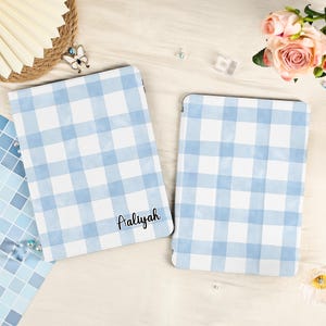 May include: Two tablet covers with a blue and white gingham design. One cover features the name "Aaliyah" in a handwritten style. The covers are displayed on a light surface, with floral and decorative accents.