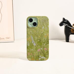 May include: A green phone case featuring a floral design with yellow, white, and pink flowers. The case has a matte finish and a camera cutout. The design is reminiscent of a meadow or garden.
