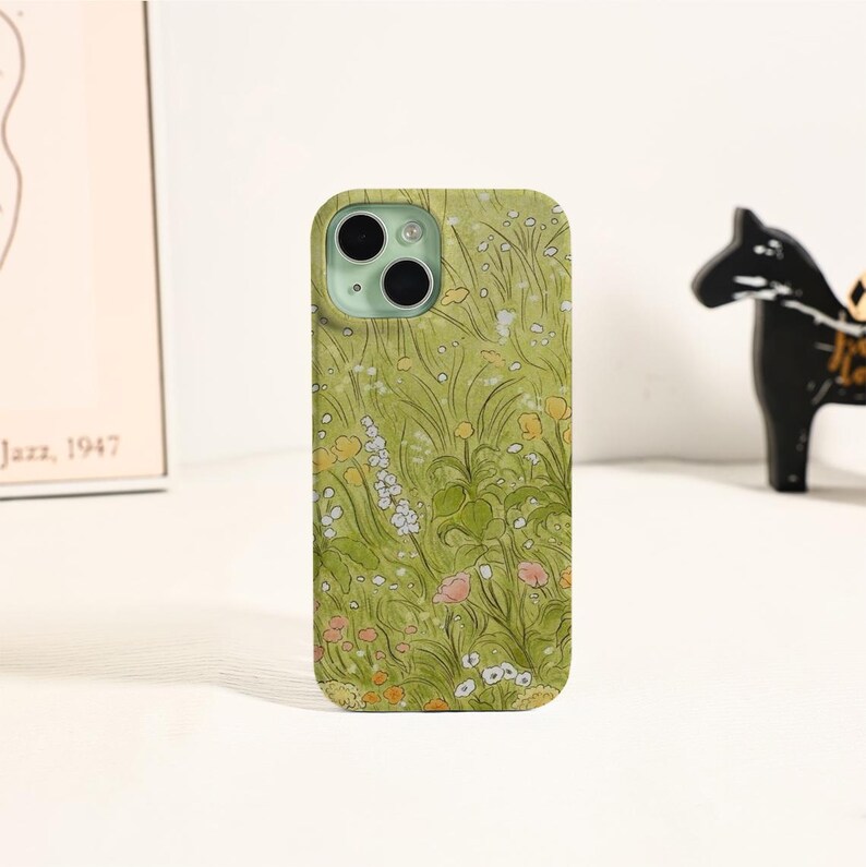May include: A green phone case featuring a floral design with yellow, white, and pink flowers. The case has a matte finish and a camera cutout. The design is reminiscent of a meadow or garden.