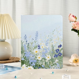 Puede incluir: Un cuaderno de tapa dura con un diseño floral. La portada presenta un fondo azul claro con ilustraciones de margaritas blancas, flores silvestres azules y follaje verde. El cuaderno es rectangular y parece estar cerrado.