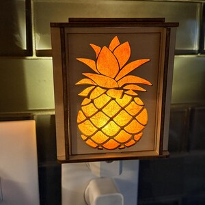 May include: A wooden night light featuring a cut-out pineapple design. The pineapple is illuminated with an orange glow, set within a rectangular frame. The night light plugs directly into a standard wall outlet.