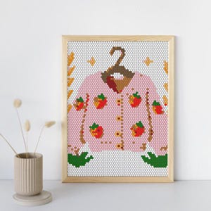 May include: A framed print featuring a pink cardigan adorned with red strawberry and yellow flower designs. The cardigan is hanging on a brown hanger. The background is a white mosaic pattern with decorative elements.