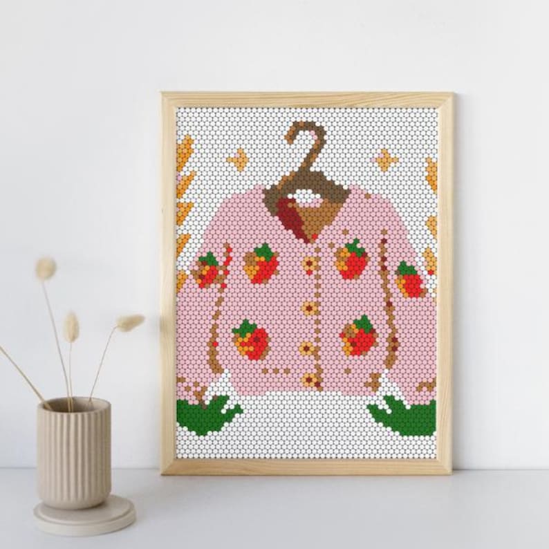 May include: A framed print featuring a pink cardigan adorned with red strawberry and yellow flower designs. The cardigan is hanging on a brown hanger. The background is a white mosaic pattern with decorative elements.