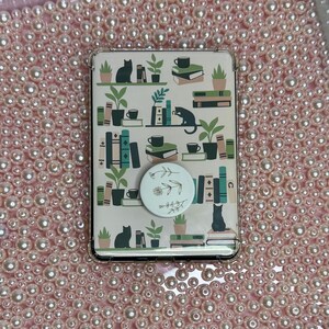 Può includere: Una custodia per telefono con sfondo color crema con un motivo di libri, piante, tazze da caffè e gatti neri. È attaccato un pop socket bianco con un design floreale. La custodia è circondata da uno sfondo di perle rosa.