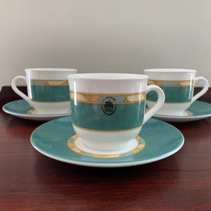 May include: Three white teacups with green and gold trim and matching saucers. The cups have a gold logo that reads "The Card Garden" with a stylized image of a garden.