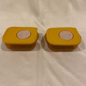 May include: Two yellow rectangular food storage containers with light tan lids. Each lid has a circular, light tan indentation in the center. The containers are on a white surface.