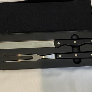 May include: A carving set presented in a black case. The set comprises a long, stainless steel carving knife and a carving fork with black handles. Each handle features three silver rivets. The case is open, displaying the tools.