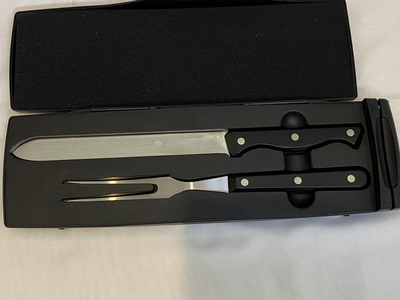 May include: A carving set presented in a black case. The set comprises a long, stainless steel carving knife and a carving fork with black handles. Each handle features three silver rivets. The case is open, displaying the tools.