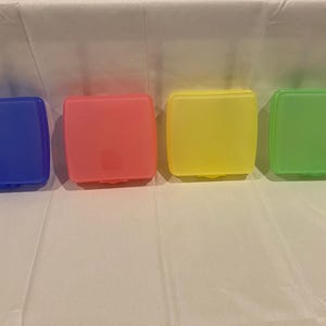 Könnte beinhalten: Vier farbige Kunststoffbehälter in Blau, Rosa, Gelb und Grün. Jeder Behälter ist rechteckig mit leicht abgerundeter Oberseite und einem kleinen Verschluss an der Vorderseite. Die Behälter sind in einer Reihe angeordnet.