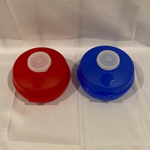 May include: Two round, plastic containers. One is red, the other is blue. Each has a white, circular lid with a small, raised center. The containers are sitting on a white surface.