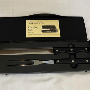 May include: A black carving set from The Pampered Chef, including a carving knife and fork, presented in a black case. The knife has a long, stainless steel blade and a black handle. The set includes a recipe card.