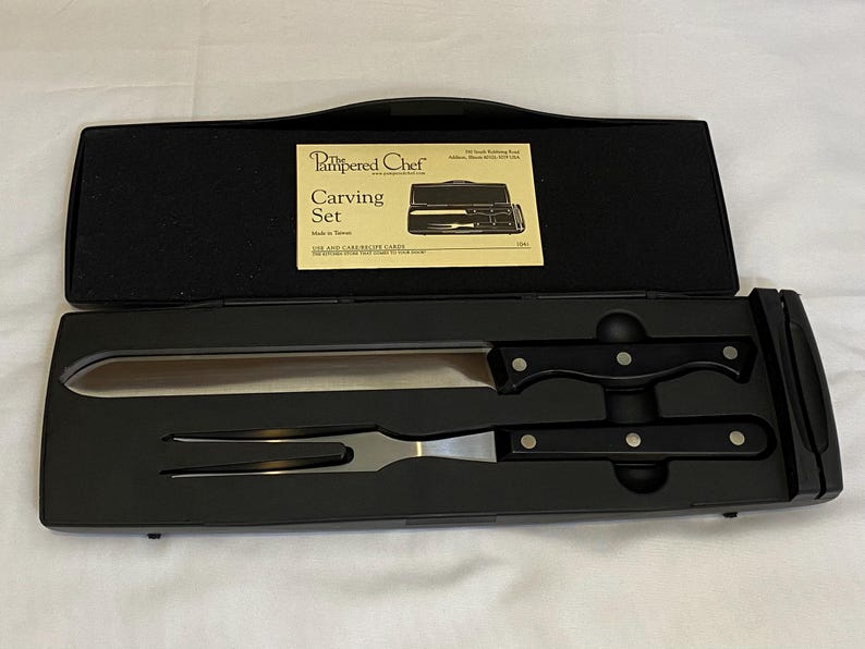 May include: A black carving set from The Pampered Chef, including a carving knife and fork, presented in a black case. The knife has a long, stainless steel blade and a black handle. The set includes a recipe card.