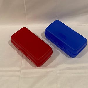 Puede incluir: Dos recipientes rectangulares de plástico. Uno es rojo brillante, el otro es azul vibrante. Ambos tienen una parte superior ligeramente redondeada y un pequeño cierre en la parte delantera. Los recipientes están sobre una superficie blanca texturizada.