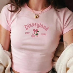 Puede incluir: Un top corto rosa claro con la palabra "Disneyland" bordada en cursiva, junto con "EST. 1955" y un gr&aacute;fico de cerezas. La camiseta tiene mangas cortas y cuello redondo.