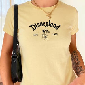 Puede incluir: Una camiseta de manga corta de color amarillo p&aacute;lido con el texto negro Disneyland, EST. 1955 y un gr&aacute;fico de Minnie Mouse. La camiseta tiene cuello redondo y se lleva con un collar de cadena dorada con colgante de coraz&oacute;n y un bolso negro.
