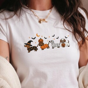 Può includere: Una t-shirt bianca a maniche corte con una grafica di quattro bassotti in costume di Halloween: da strega, dentro una zucca, da mummia e da fantasma con una zucca, circondati da pipistrelli, stelle e una falce di luna.
