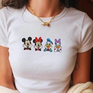 May include: White t-shirt featuring a colorful graphic of Mickey Mouse, Minnie Mouse, Donald Duck, and Daisy Duck. The cartoon characters are in a row, each with a unique pose and outfit. The shirt has a classic crew neck design.