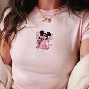 K&ouml;nnte beinhalten: Hellrosa T-Shirt mit einer Cartoonfigur mit schwarzen Mauseohren, einem schwarzen Stirnband und einem kleinen Teddyb&auml;ren. Die Figur ist rosa und lila, mit gro&szlig;en Ohren und einem niedlichen Ausdruck. Das T-Shirt hat einen runden Ausschnitt.