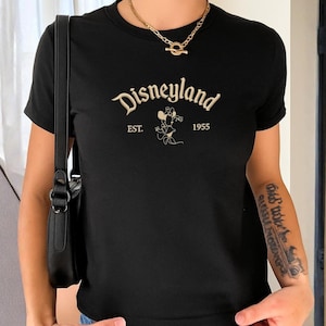 Puede incluir: Una camiseta negra de manga corta que presenta la palabra Disneyland en una fuente cursiva de color crema, con el texto EST. 1955 y una ilustraci&oacute;n de l&iacute;nea de Minnie Mouse centrada debajo del t&iacute;tulo principal en un tono crema a juego.