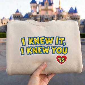 May include: A beige crewneck sweatshirt featuring embroidered text that reads I KNEW IT, I KNEW YOU in yellow with blue outlines. Below the text is a red heart shape containing the yellow embroidered letters TS with a blue outline.