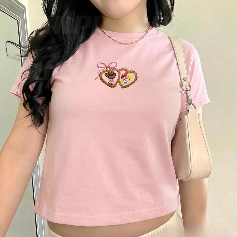 May include: A light pink t-shirt featuring an embroidered design of two heart-shaped frames. One frame contains a cowboy, the other a cowgirl. The shirt has short sleeves and a crew neck.