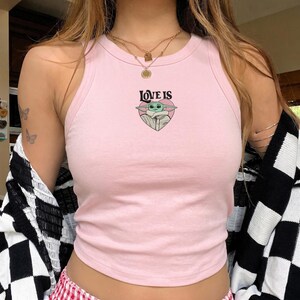 May include: A light pink sleeveless tank top featuring a central graphic of a green creature inside a pink heart with the text LOVE IS printed in black above the heart. The garment has a ribbed crew neckline and a fitted silhouette.