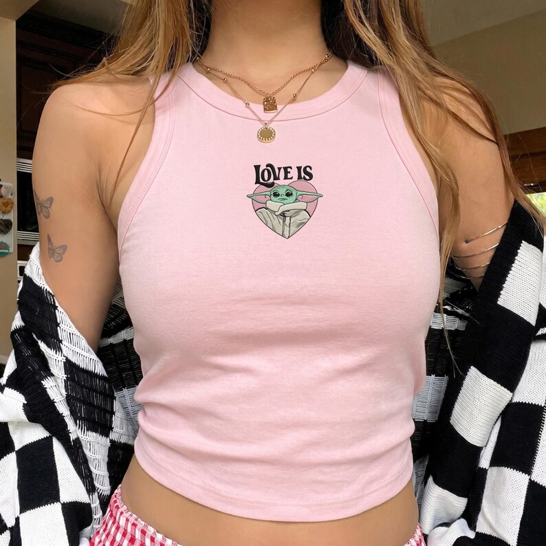May include: A light pink sleeveless tank top featuring a central graphic of a green creature inside a pink heart with the text LOVE IS printed in black above the heart. The garment has a ribbed crew neckline and a fitted silhouette.