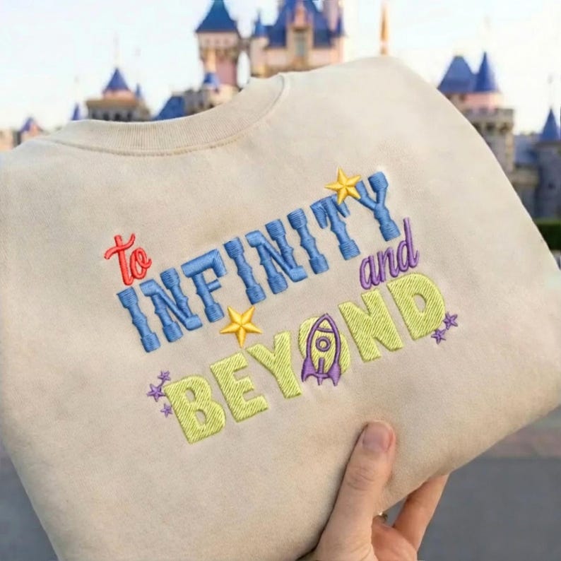 May include: A beige crewneck sweatshirt featuring embroidered text that reads To INFINITY and BEYOND. The word To is in red, INFINITY is in blue, and BEYOND is in lime green. The design includes yellow stars, small purple stars, and a purple rocket ship.