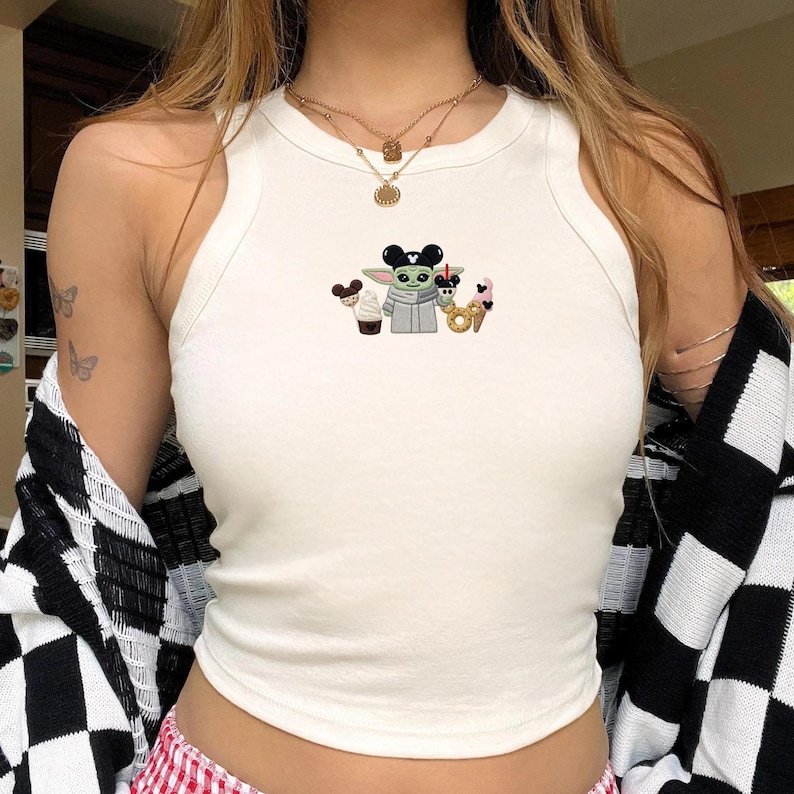 May include: A cream-colored sleeveless tank top featuring a central embroidered graphic of a green alien character wearing black mouse ears, holding a drink, and surrounded by a cupcake, a pretzel, and an ice cream cone with mouse-shaped details.