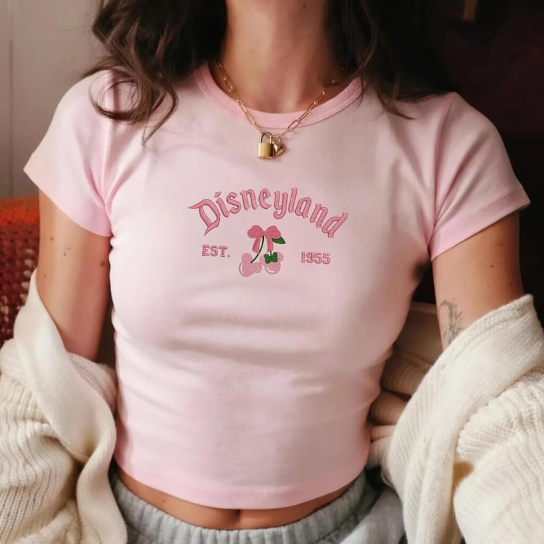 Puede incluir: Un top corto rosa claro con la palabra "Disneyland" bordada en cursiva, junto con "EST. 1955" y un gr&aacute;fico de cerezas. La camiseta tiene mangas cortas y cuello redondo.
