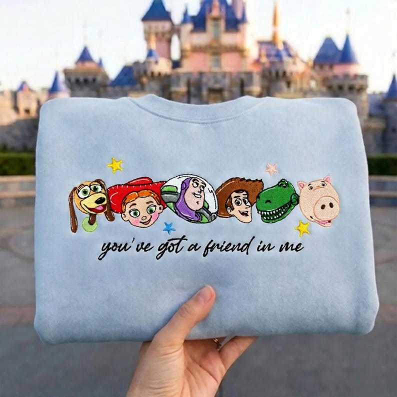 May include: A light blue crewneck sweatshirt featuring embroidered character faces from Toy Story, including Slinky Dog, Jessie, Buzz Lightyear, Woody, Rex, and Hamm, with the black cursive text "you've got a friend in me" and small yellow stars.