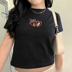 May include: Black t-shirt with a crew neck featuring embroidered heart-shaped frames with cartoon characters. The frames are connected by a pink bow. A small, light-colored purse hangs from the shoulder.