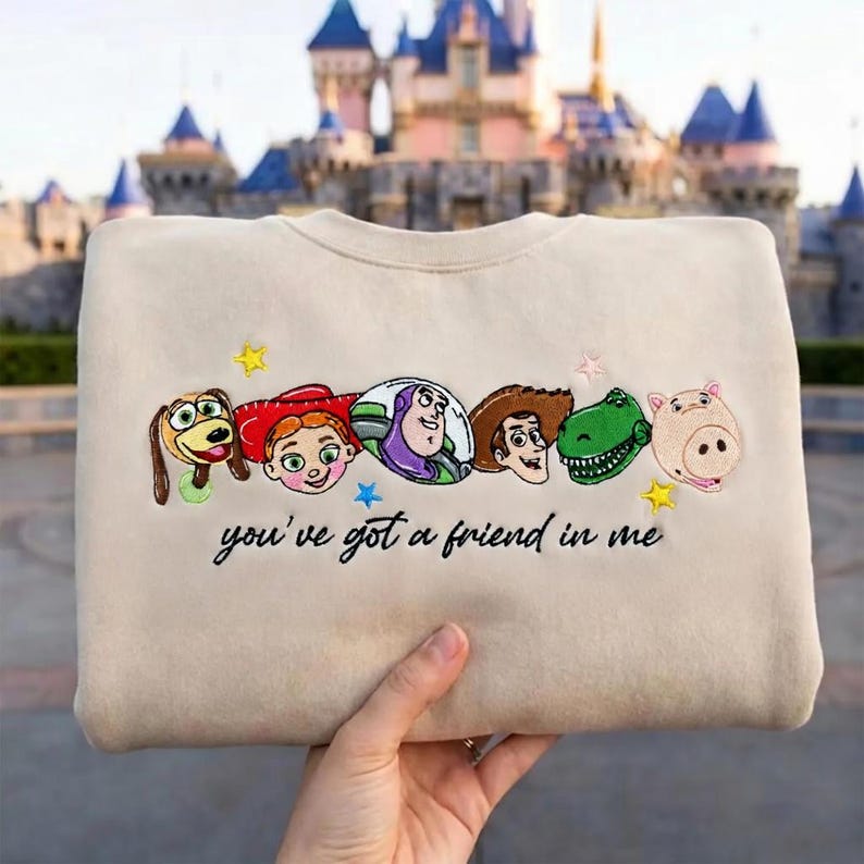 May include: A beige crewneck sweatshirt featuring embroidered character faces from Toy Story, including Slinky Dog, Jessie, Buzz Lightyear, Woody, Rex, and Hamm, with the embroidered text "you've got a friend in me" and small yellow stars.