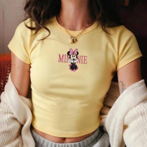 Może przedstawiać: Ż&oacute;łty top z nadrukiem Myszki Minnie. Grafika zawiera napis "Minnie" w r&oacute;żowych literach nad kresk&oacute;wkowym wizerunkiem Myszki Minnie w r&oacute;żowej kokardce i sukience.